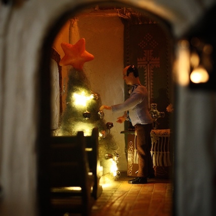 Jouluviikko. Kuva nukkekotikokoisen kappelin ovelta. Alttarin edessä Suntio kohentamassa joulukuusta. 