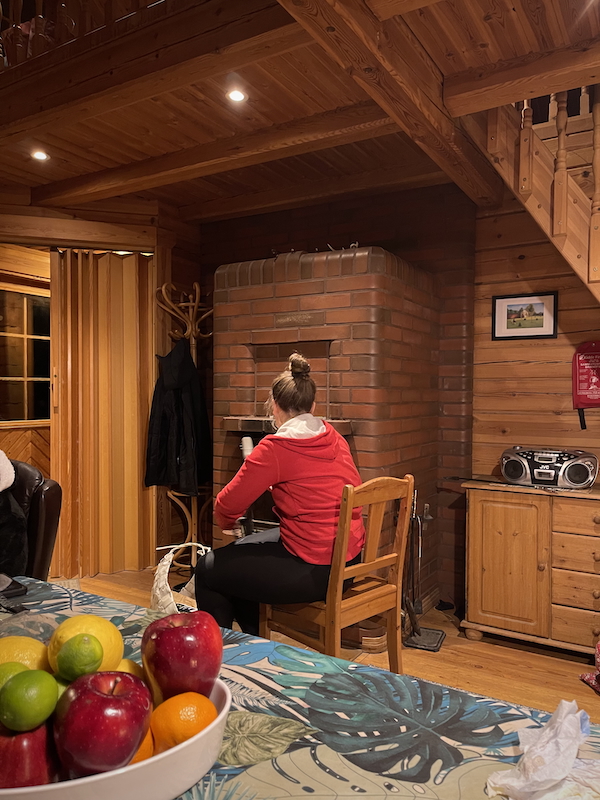 Joulukuun valoja ja varjoja. Kuvassa lomamökki sisältä. Keskellä kuvaa takka ja nainen joka katsoo takkaan päin.