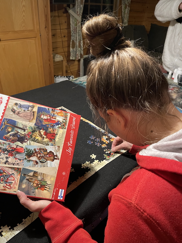 Joulukuun valoja ja varjoja. Kuvassa nainen kokoaa palapeliä. Palapelin kuvituksena ovat vanhat joulukortit.