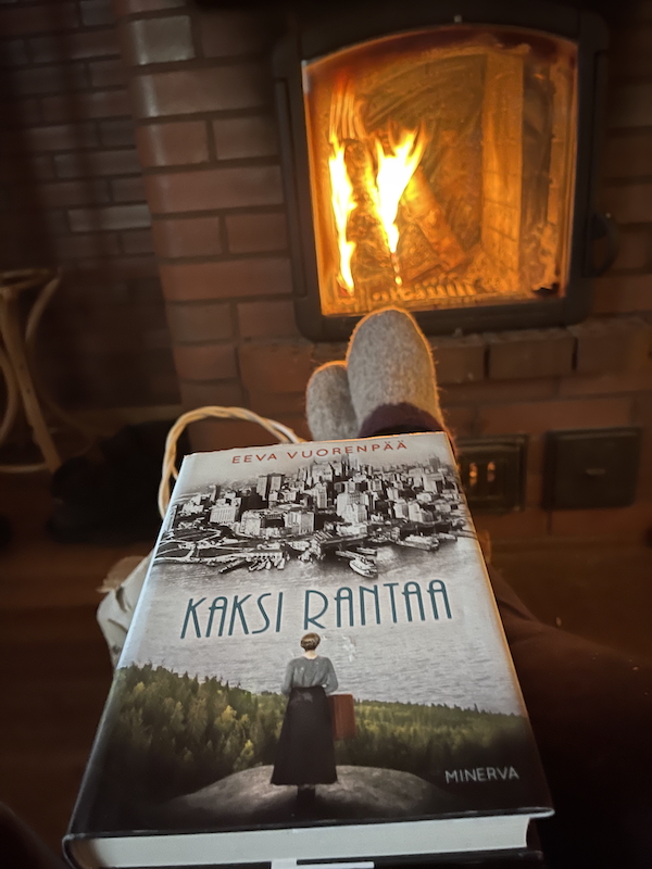 Joulukuun valoja ja varjoja. Kuvassa naisenjalat, kirja ja takka jossa palaa tuli.