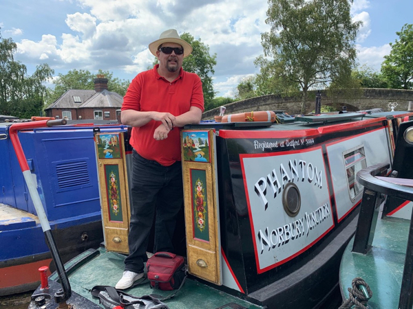 Kolme asiaa elämästä. Narrowboat, jonka perässä seisoo mies punaisessa paidassa.